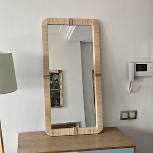 Può includere: Uno specchio rettangolare con una cornice intrecciata naturale. Lo specchio è appeso a una parete bianca sopra una cassettiera in legno.