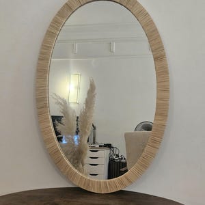 May include: Oval mirror with a woven, natural-toned frame. The mirror reflects a room with a white wall, a lamp, and a dark wooden table. The frame's texture adds a rustic, bohemian aesthetic.