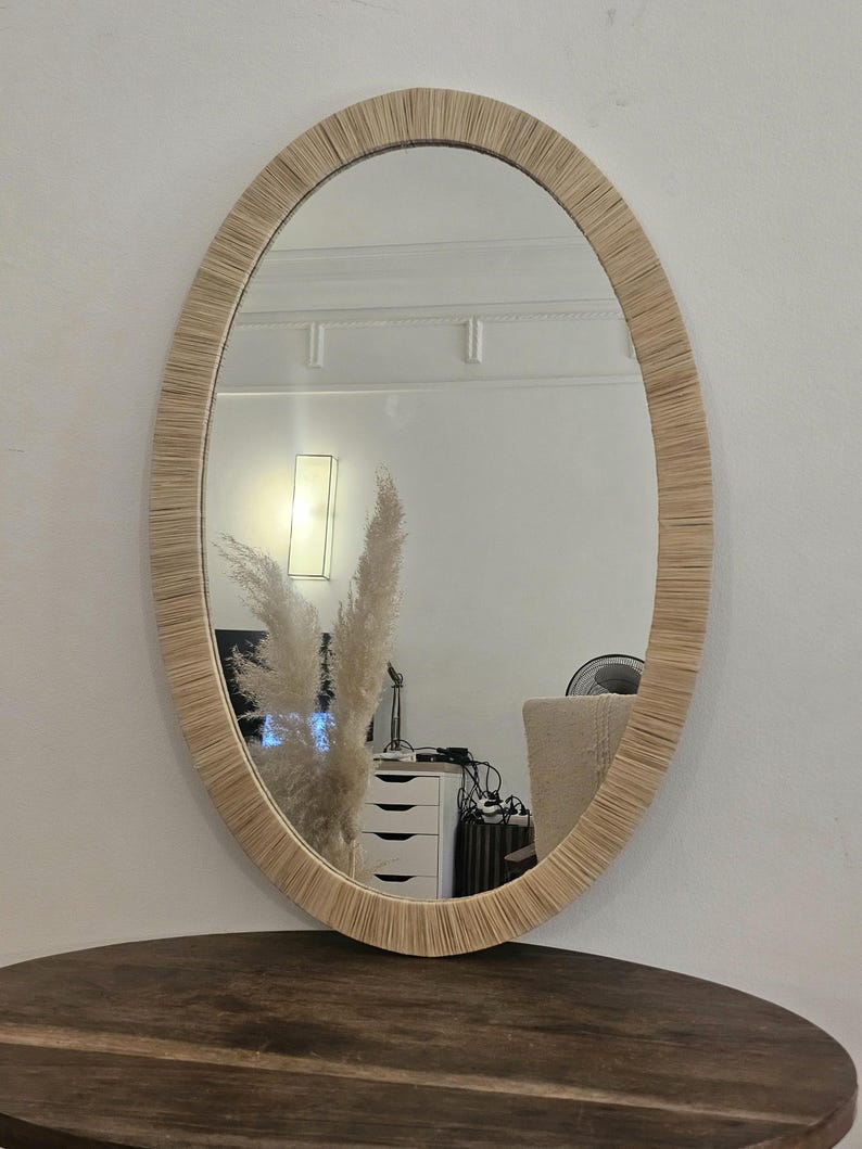 May include: Oval mirror with a woven, natural-toned frame. The mirror reflects a room with a white wall, a lamp, and a dark wooden table. The frame's texture adds a rustic, bohemian aesthetic.