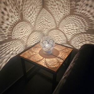 Puede incluir: Una l&aacute;mpara de mesa de metal plateado con una pantalla redonda y estampada proyecta intrincadas sombras geom&eacute;tricas en una mesa de madera y una pared. La l&aacute;mpara est&aacute; sobre una mesa peque&ntilde;a, cuadrada, de metal negro.
