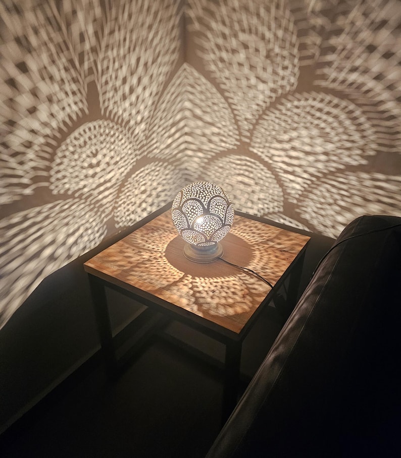 Puede incluir: Una l&aacute;mpara de mesa de metal plateado con una pantalla redonda y estampada proyecta intrincadas sombras geom&eacute;tricas en una mesa de madera y una pared. La l&aacute;mpara est&aacute; sobre una mesa peque&ntilde;a, cuadrada, de metal negro.