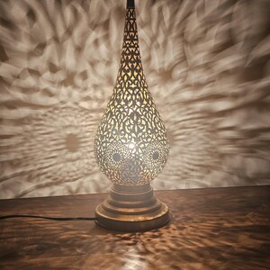 May include: A decorative brass table lamp with an intricate, openwork design. The lamp has a teardrop-shaped shade and a tall, slender top. The light casts a patterned shadow on the wall behind it. The lamp is on a wooden surface.