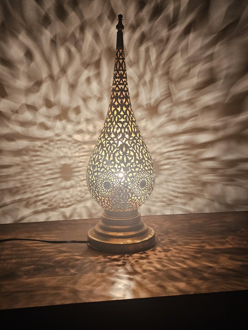 May include: A decorative brass table lamp with an intricate, openwork design. The lamp has a teardrop-shaped shade and a tall, slender top. The light casts a patterned shadow on the wall behind it. The lamp is on a wooden surface.