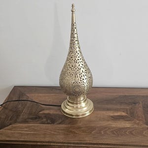 May include: A gold-colored metal table lamp with a pierced, teardrop-shaped shade and a round base. The lamp has an elongated, pointed top and intricate cut-out patterns. The lamp is plugged into a dark wooden surface.