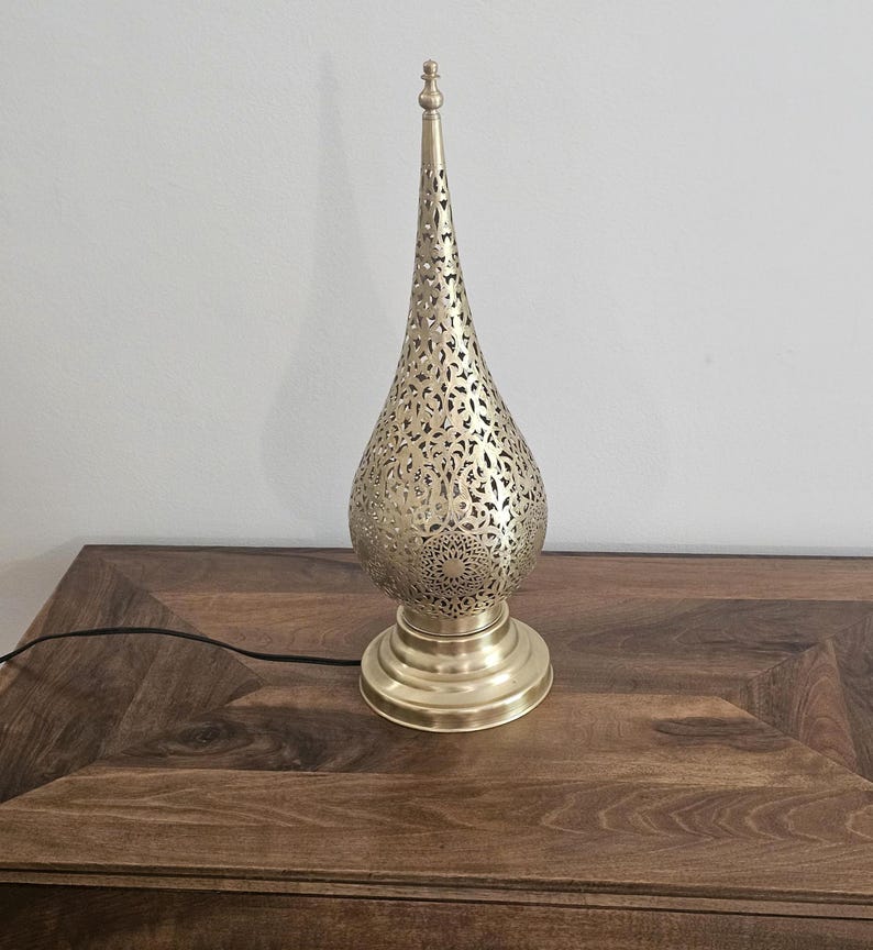 May include: A gold-colored metal table lamp with a pierced, teardrop-shaped shade and a round base. The lamp has an elongated, pointed top and intricate cut-out patterns. The lamp is plugged into a dark wooden surface.