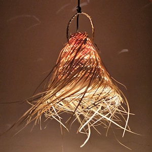 Puede incluir: Una lámpara colgante tejida hecha de materiales naturales, colgada de un cordón negro. La lámpara tiene forma de cono y es de color marrón claro.