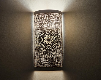 May include: A white metal wall sconce with a circular, intricate design. The sconce is illuminated, casting a warm glow.