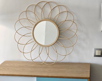 May include: A round, wooden, decorative mirror with a flower-shaped frame. The mirror has a circular, reflective surface. The frame is made of thin, woven strips of wood.