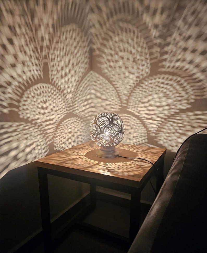Puede incluir: Una l&aacute;mpara de mesa de metal plateado con una pantalla redonda y estampada proyecta intrincadas sombras geom&eacute;tricas sobre una mesa de madera y un sof&aacute; cercano.