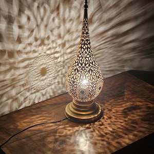 May include: A decorative brass lamp with a pierced, teardrop-shaped shade. The lamp sits on a round base and has a long, pointed top. The lamp casts intricate patterns of light on the surrounding surface.