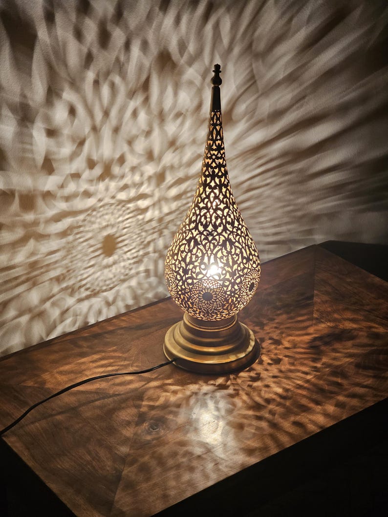 May include: A decorative brass lamp with a pierced, teardrop-shaped shade. The lamp sits on a round base and has a long, pointed top. The lamp casts intricate patterns of light on the surrounding surface.