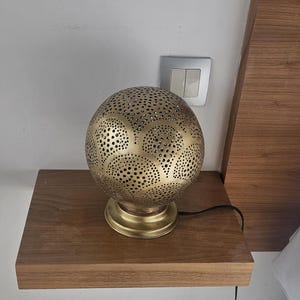 Puede incluir: Una l&aacute;mpara de mesa de metal dorado con una pantalla redonda perforada. La pantalla tiene un patr&oacute;n geom&eacute;trico de c&iacute;rculos y &oacute;valos. La l&aacute;mpara est&aacute; sobre una mesa de madera.