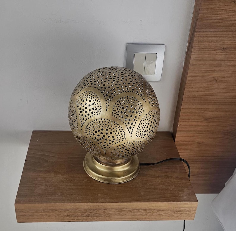 Puede incluir: Una l&aacute;mpara de mesa de metal dorado con una pantalla redonda perforada. La pantalla tiene un patr&oacute;n geom&eacute;trico de c&iacute;rculos y &oacute;valos. La l&aacute;mpara est&aacute; sobre una mesa de madera.