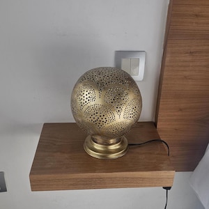 Puede incluir: Una l&aacute;mpara de mesa de metal dorado con una pantalla redonda perforada. La l&aacute;mpara est&aacute; sobre una repisa de madera.