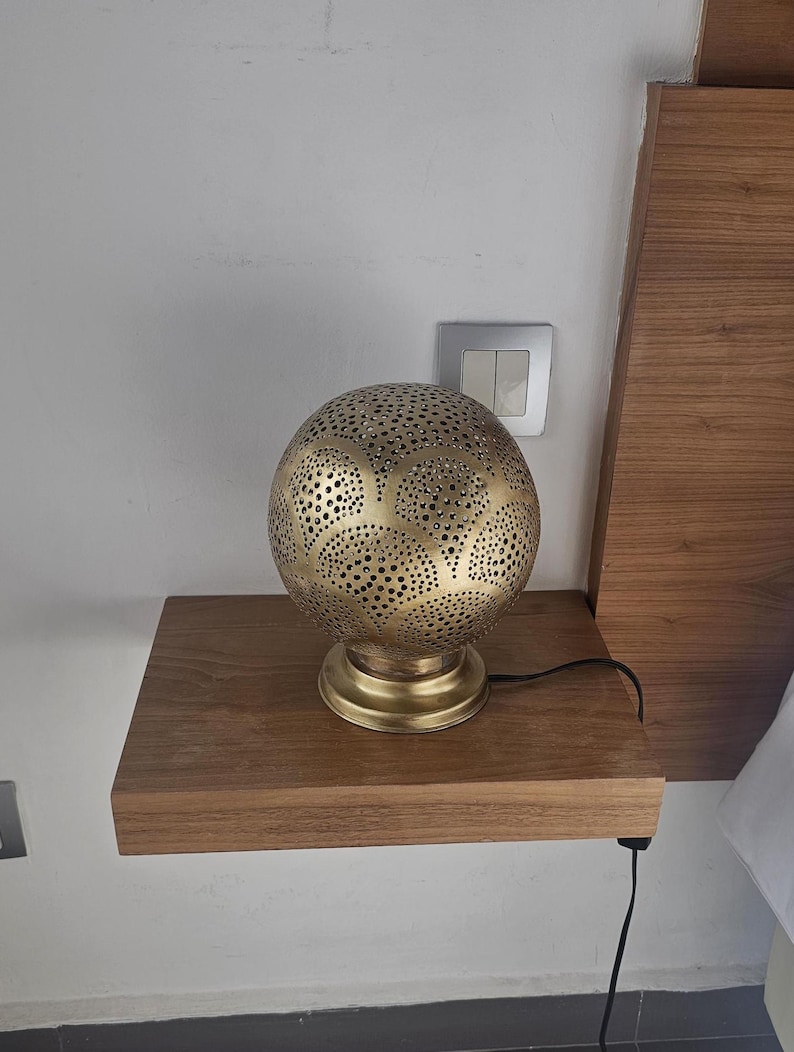 Puede incluir: Una l&aacute;mpara de mesa de metal dorado con una pantalla redonda perforada. La l&aacute;mpara est&aacute; sobre una repisa de madera.