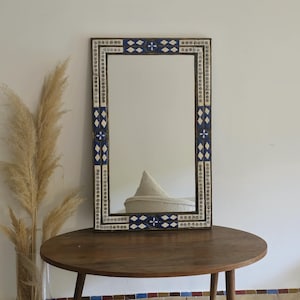 May include: A decorative mirror with a blue and white mosaic frame. The frame features geometric patterns and is set against a white wall.
