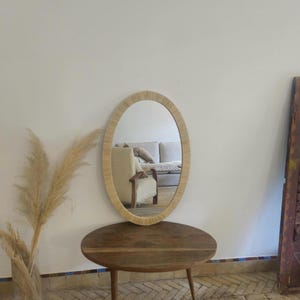 May include: Oval mirror with a woven, light-colored frame. The mirror is displayed on a round, wooden table with three legs. The reflection shows a glimpse of a living room with a sofa and chair.