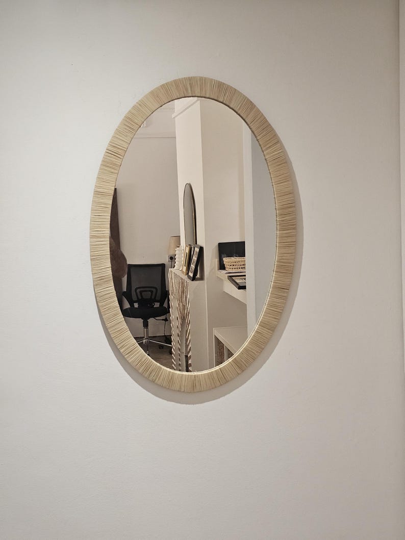 May include: Oval mirror with a woven, light-colored frame. The mirror is hanging on a white wall, reflecting a room with a black chair and other furnishings. The frame has a textured appearance.