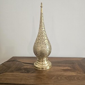 May include: A gold-colored metal table lamp with a teardrop-shaped shade featuring intricate cut-out patterns. The lamp has a round base and a tall, pointed top. The lamp is plugged into a wooden table.