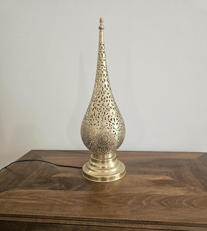 May include: A gold-colored metal table lamp with a teardrop-shaped shade featuring intricate cut-out patterns. The lamp has a round base and a tall, pointed top. The lamp is plugged into a wooden table.