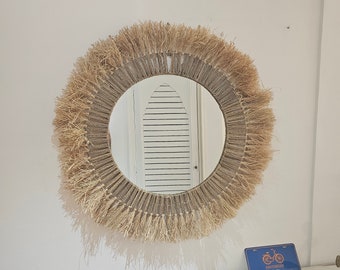 May include: A round mirror with a natural fiber frame. The frame is made of woven straw with a fringe around the edge. The mirror is reflecting a white door with vertical slats.