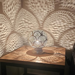 Puede incluir: Una l&aacute;mpara de mesa plateada con una pantalla redonda y estampada proyecta intrincadas sombras geom&eacute;tricas sobre una mesa de madera. La l&aacute;mpara est&aacute; en un cable negro y tiene una base blanca.