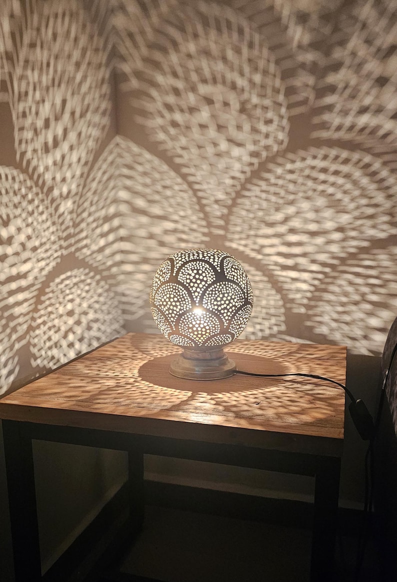 Puede incluir: Una l&aacute;mpara de mesa plateada con una pantalla redonda y estampada proyecta intrincadas sombras geom&eacute;tricas sobre una mesa de madera. La l&aacute;mpara est&aacute; en un cable negro y tiene una base blanca.