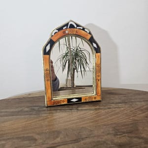 Op de afbeelding: Een kleine, gebogen spiegel met een decoratief frame. Het frame is gemaakt van hout en heeft een zwart, wit en oranje geometrisch patroon. De spiegel reflecteert een plant in een pot.