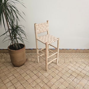 Op de afbeelding: Een lichte houten barstoel met een gevlochten leren zitting en rugleuning. De stoel staat in een kamer met een betegelde vloer en een plant in een pot.