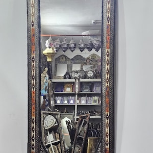 May include: A tall, rectangular mirror with an arched top. The frame is black with intricate patterns in orange, red, and silver. The mirror reflects a shop interior with other mirrors and decorative items. The mirror is a decorative home decor item.