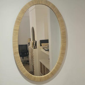 May include: Oval mirror with a natural woven frame. The mirror reflects a room with a black office chair, a white shelf, and other decorative items. The frame has a light beige color.
