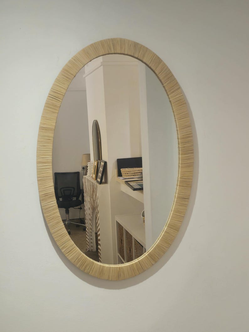 May include: Oval mirror with a natural woven frame. The mirror reflects a room with a black office chair, a white shelf, and other decorative items. The frame has a light beige color.
