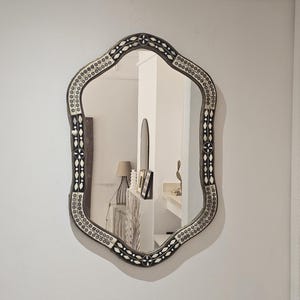 Può includere: Uno specchio decorativo ornato, con una forma unica a smerlo. La cornice scura presenta un motivo dettagliato di piccoli accenti rotondi e ovali bianchi e argentati. Lo specchio è appeso a una parete bianca.