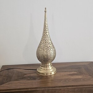 May include: A gold-colored metal table lamp with a teardrop-shaped shade featuring intricate cut-out patterns. The lamp has a round base and a long, pointed top. A black power cord extends from the base.