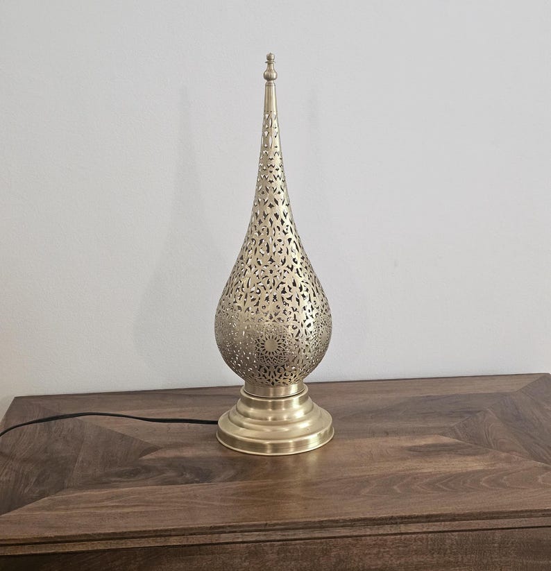 May include: A gold-colored metal table lamp with a teardrop-shaped shade featuring intricate cut-out patterns. The lamp has a round base and a long, pointed top. A black power cord extends from the base.