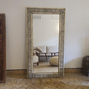 Pode incluir: Um espelho retangular grande com uma moldura ornamentada de cor creme. A moldura apresenta um padrão detalhado e repetitivo em metal prateado. O espelho reflete uma sala com um sofá branco, uma mesa de madeira e um pufe tecido. O espelho está sobre um chão ladrilhado.