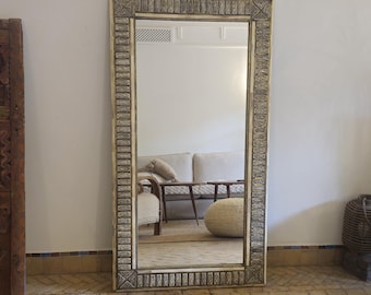 May include: A large rectangular mirror with an ornate, cream-colored frame. The frame features a detailed, repeating pattern in a silver-tone metal. The mirror reflects a room with a white sofa, a wooden table, and a woven ottoman. The mirror is standing on a tiled floor.