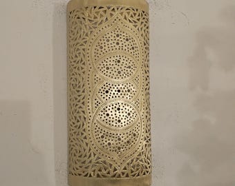 May include: A cylindrical, gold-colored wall sconce with intricate cut-out patterns. The design features geometric shapes and circular perforations, creating a decorative lighting element. The sconce is made of metal and has a warm, inviting appearance.
