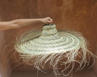 Puede incluir: Una gran pantalla de lámpara de paja tejida con un ala ancha y un borde con flecos. La pantalla de la lámpara está sujeta por la mano de una persona. El color natural de la paja crea una estética cálida. La pantalla de la lámpara está diseñada para colgar del techo.