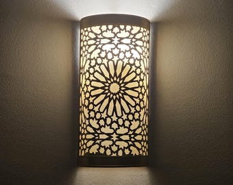 May include: A white wall sconce with a geometric pattern and a light bulb illuminating the intricate design.