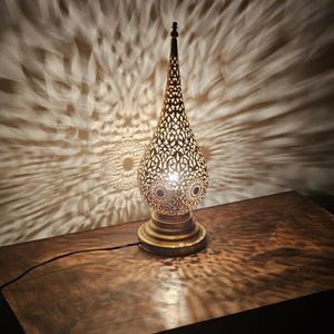 May include: A decorative brass table lamp with an intricate, openwork design. The lamp has a teardrop shape and sits on a round base. The light casts a patterned shadow on the wooden surface below. The lamp is plugged in.