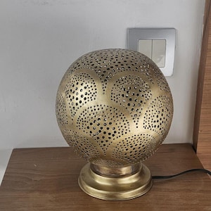 Puede incluir: Una l&aacute;mpara de mesa de metal dorado con una pantalla redonda perforada. La pantalla tiene un patr&oacute;n decorativo de c&iacute;rculos y hojas. La l&aacute;mpara est&aacute; sobre una mesa de madera.