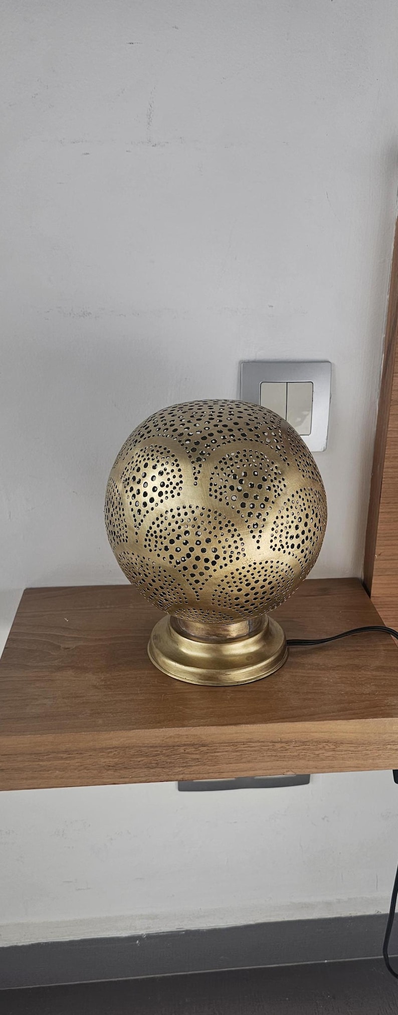 Puede incluir: Una l&aacute;mpara de mesa de metal dorado con una pantalla redonda perforada. La pantalla tiene un patr&oacute;n decorativo de c&iacute;rculos y hojas. La l&aacute;mpara est&aacute; sobre una mesa de madera.