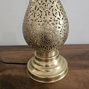 May include: A gold-colored, decorative table lamp with an intricate, openwork design. The lamp has a teardrop-shaped shade with floral and geometric patterns. The base is round and tiered. A black cord extends from the base.