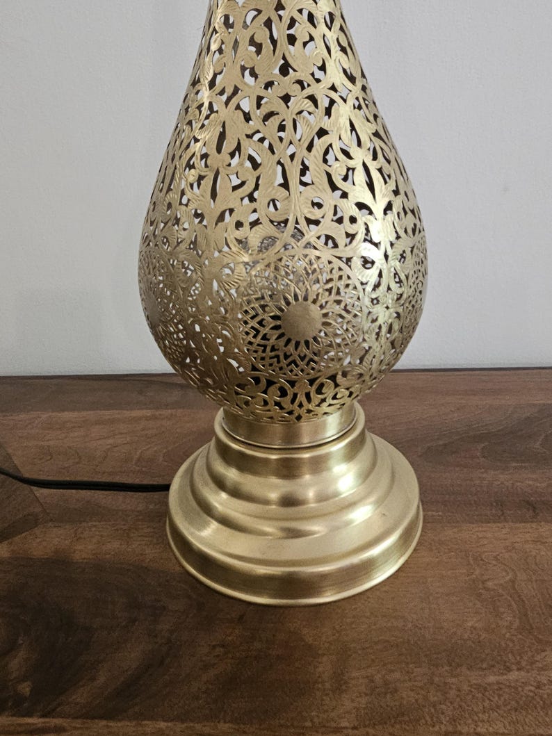 May include: A gold-colored, decorative table lamp with an intricate, openwork design. The lamp has a teardrop-shaped shade with floral and geometric patterns. The base is round and tiered. A black cord extends from the base.