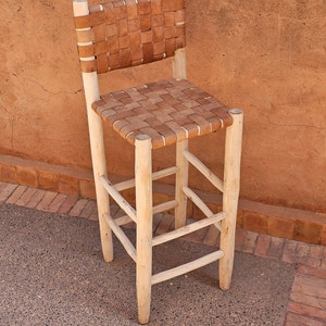 Könnte beinhalten: Ein Barhocker aus Holz mit einem geflochtenen Ledersitz und -lehne. Der Hocker hat eine helle Holzoptik und steht auf einem Betonboden.