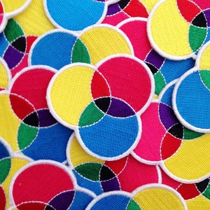 Puede incluir: Primer plano de parches de tela coloridos con un diseño circular. Los parches están hechos de una tela suave y texturizada y presentan círculos superpuestos de color amarillo, rojo, azul, verde y negro.