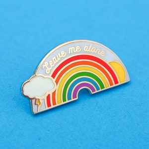 Puede incluir: Un pin de esmalte dorado con un arcoíris, una nube y un rayo. El arcoíris tiene un fondo blanco con el texto "Leave me alone" en letra cursiva.