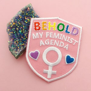 Peut inclure: Patch en tissu rose avec des coutures blanches et un texte brodé arc-en-ciel qui dit "Behold My Feminist Agenda". Un symbole féminin blanc et deux cœurs roses et bleus sont également brodés sur le patch.