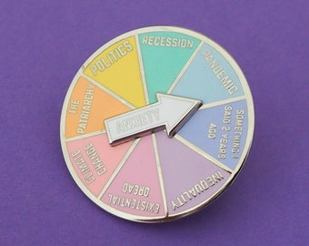 Op de afbeelding: Een zilveren pin met een kleurrijk wiel waarop de woorden "INEQUALITY", "PANDEMIC", "SOMETHING! SAID 2 YEARS AGO", "DREAD", "EXISTENTIAL", "THE PATRIARCHY", "POLITICS", "RECESSION", "CLIMATE CHANGE" staan. Het wiel wijst naar het woord "ANXIETY".